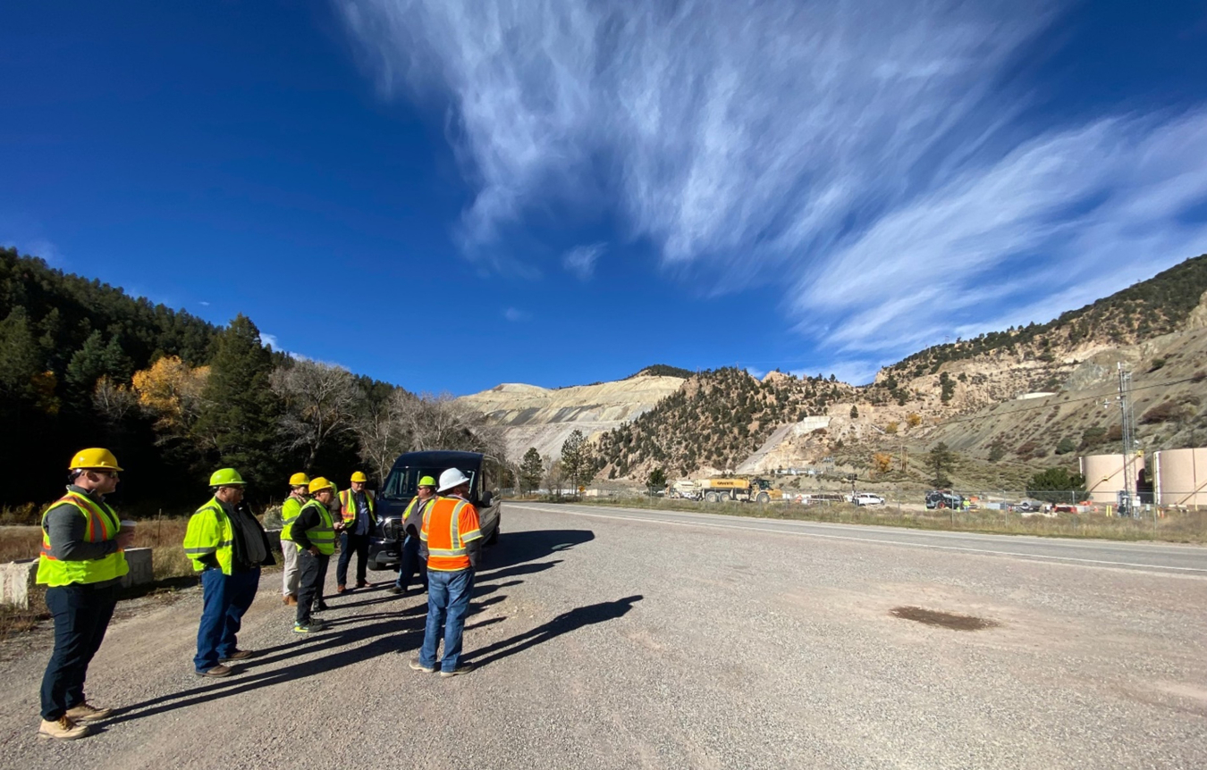 A group of seven people wearing hard hats and hi-visibility vest by a mountain road and mine.