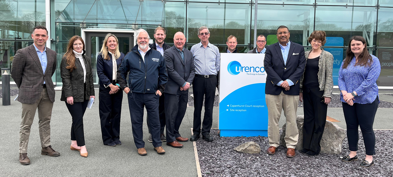 A dozen people pose for a photo in front of a building. They are next to a blue and white sign that says Urenco.