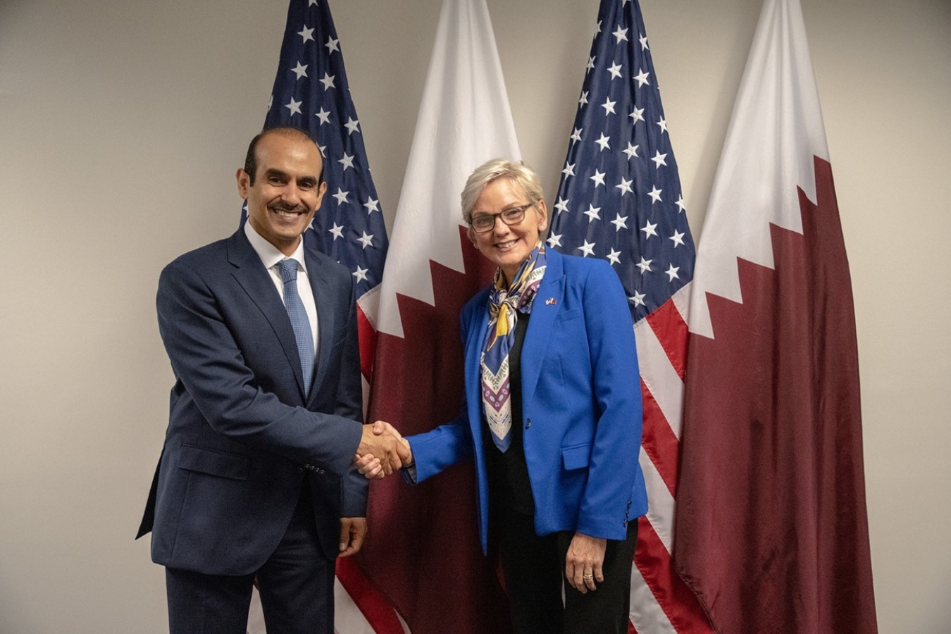 U.S. Secretary of Energy Jennifer Granholm stands in front of flags with Minister of State Al Kaabi from Qatar while shaking hands.
