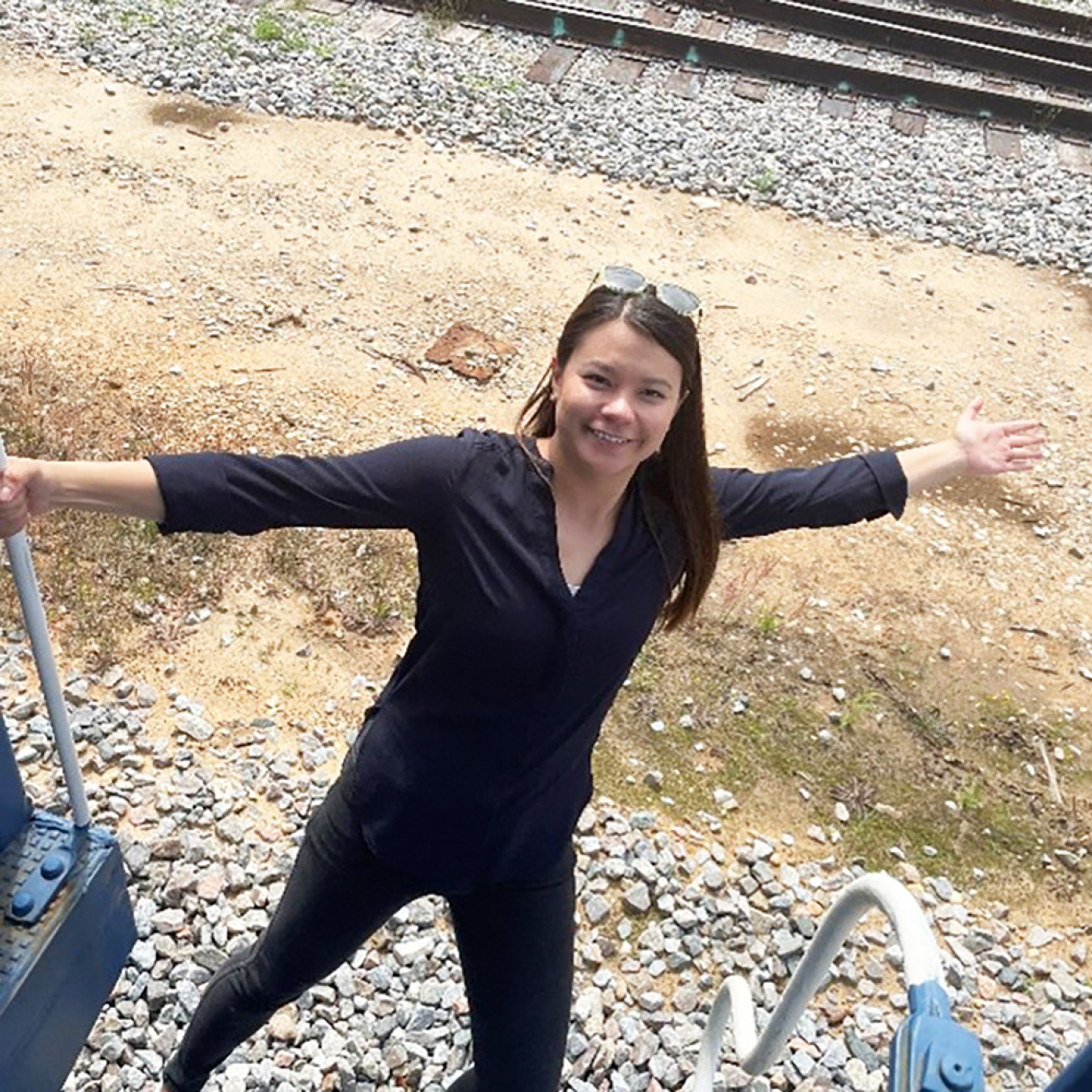 A photo of Alexa Erickson. She is smiling and hanging of the stairway to a train. Railroad tracks are behind her.