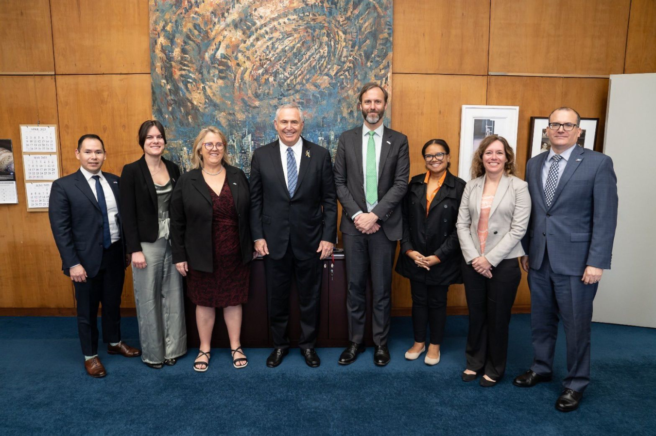 Department Net Zero World representatives, the United States Ambassador to Argentina. and the Ambassador's staff pose for a photo in the Ambassador's office in front of a light brown wall.