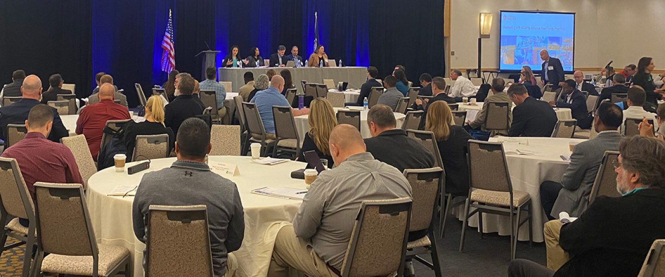 Attendees of the federal utilities partnership working group 2023 seminar sitting at tables listening to a speaker panel at the front of the room.