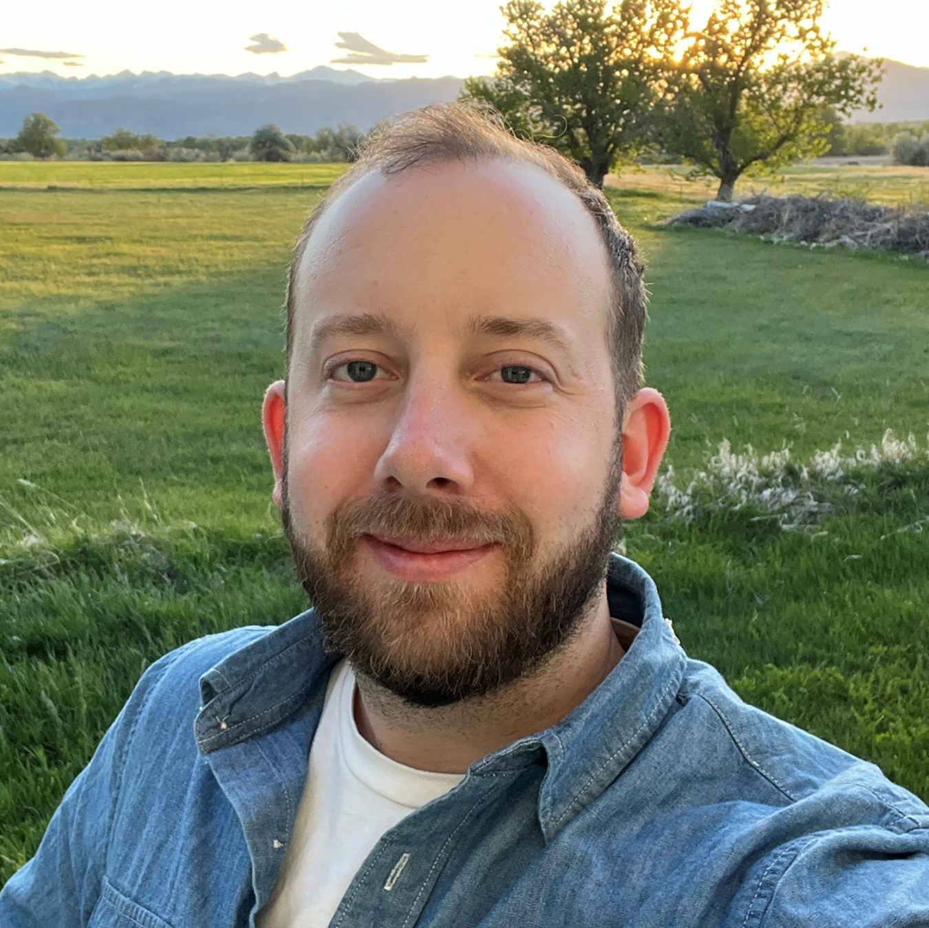 A portrait of Jesse Altum. He is dressed casually and outside with trees and a sunset or sunrise in the background.