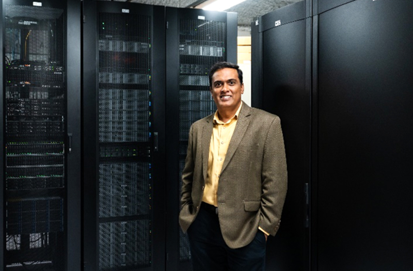 Photo of Giri Prakash (a South Asian man in a brown suit jacket and black pants) standing in front of black supercomputer cabinets