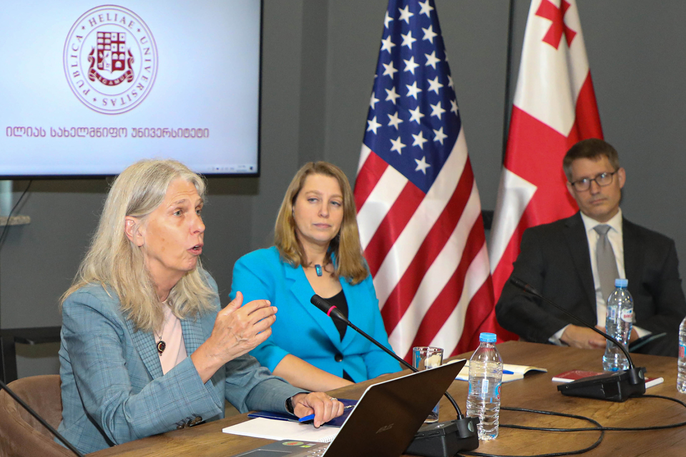Administrator Hruby speaks while she and Hinderstein are seated at a desk. Large U.S. and Georgian flags are next to them.