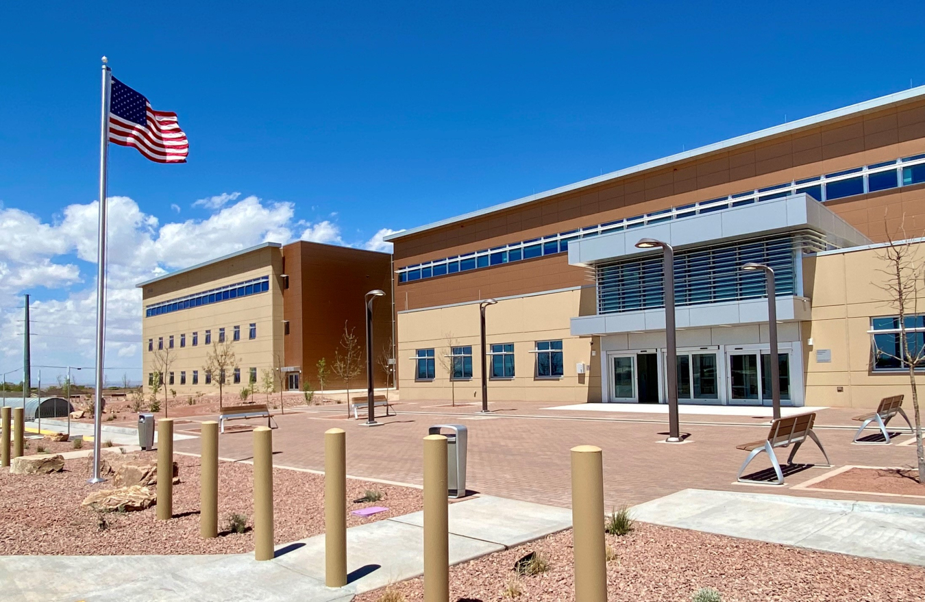 A large building that is tan is juxtaposed against a bright blue sky. An American flag flies on a pole nearby.