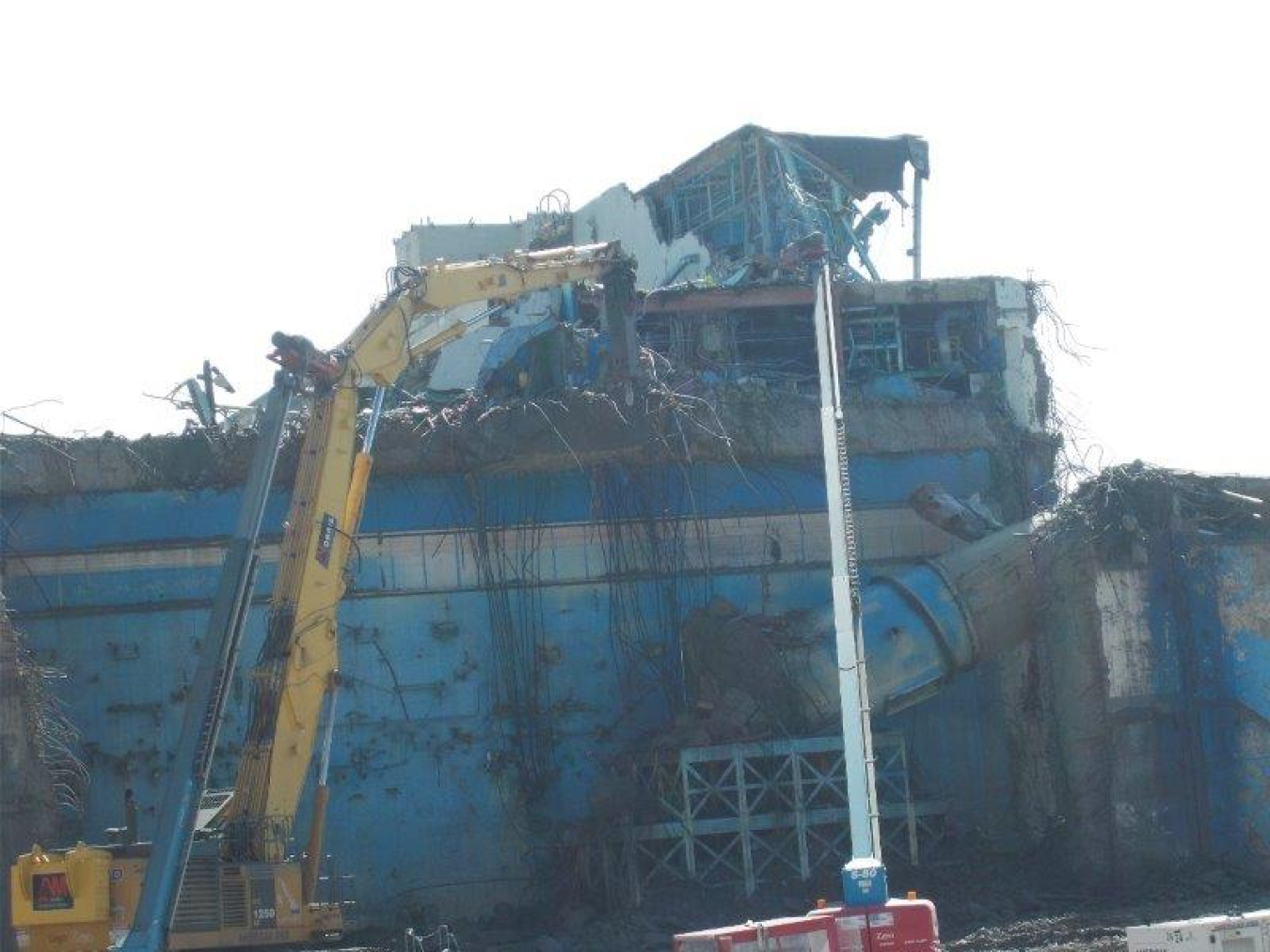 An operator at the West Valley Demonstration Project uses a heavy-duty excavator with a hydraulic hammer attachment to safely remove the remaining section of the Main Plant Process Building’s ventilation stack, weighing more than 125,000 pounds, pictured at top-right.