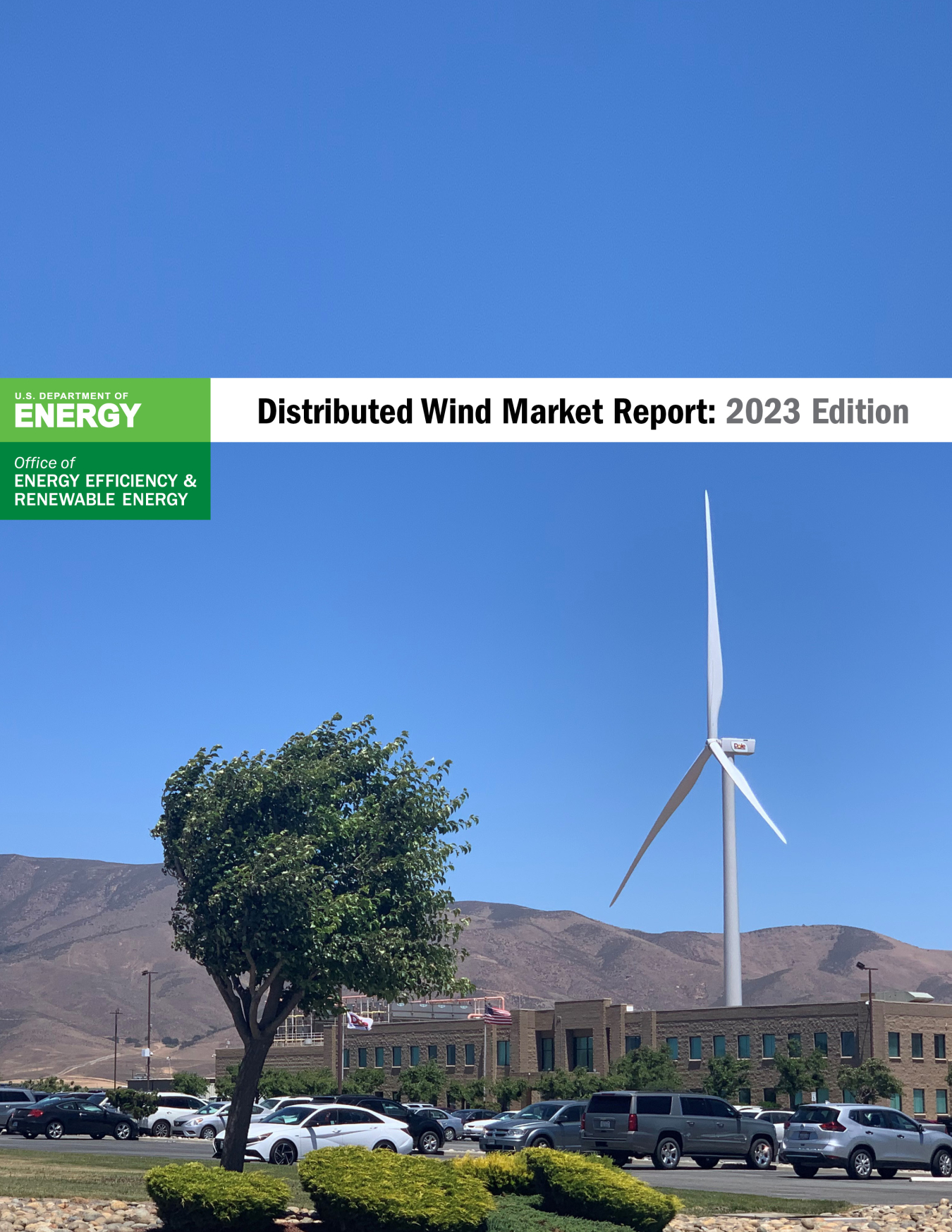 A photo of a wind turbine behind a large building and wind-blown tree overlain by the U.S. Department of Energy Wind Energy Technologies Office logo and the title Distributed Wind Market Report: 2023 Edition