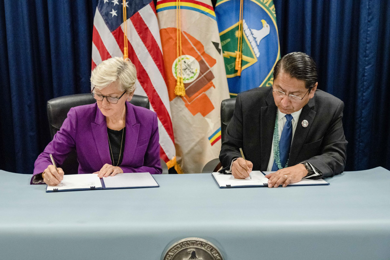 Two politicians sign documents in front of flags.