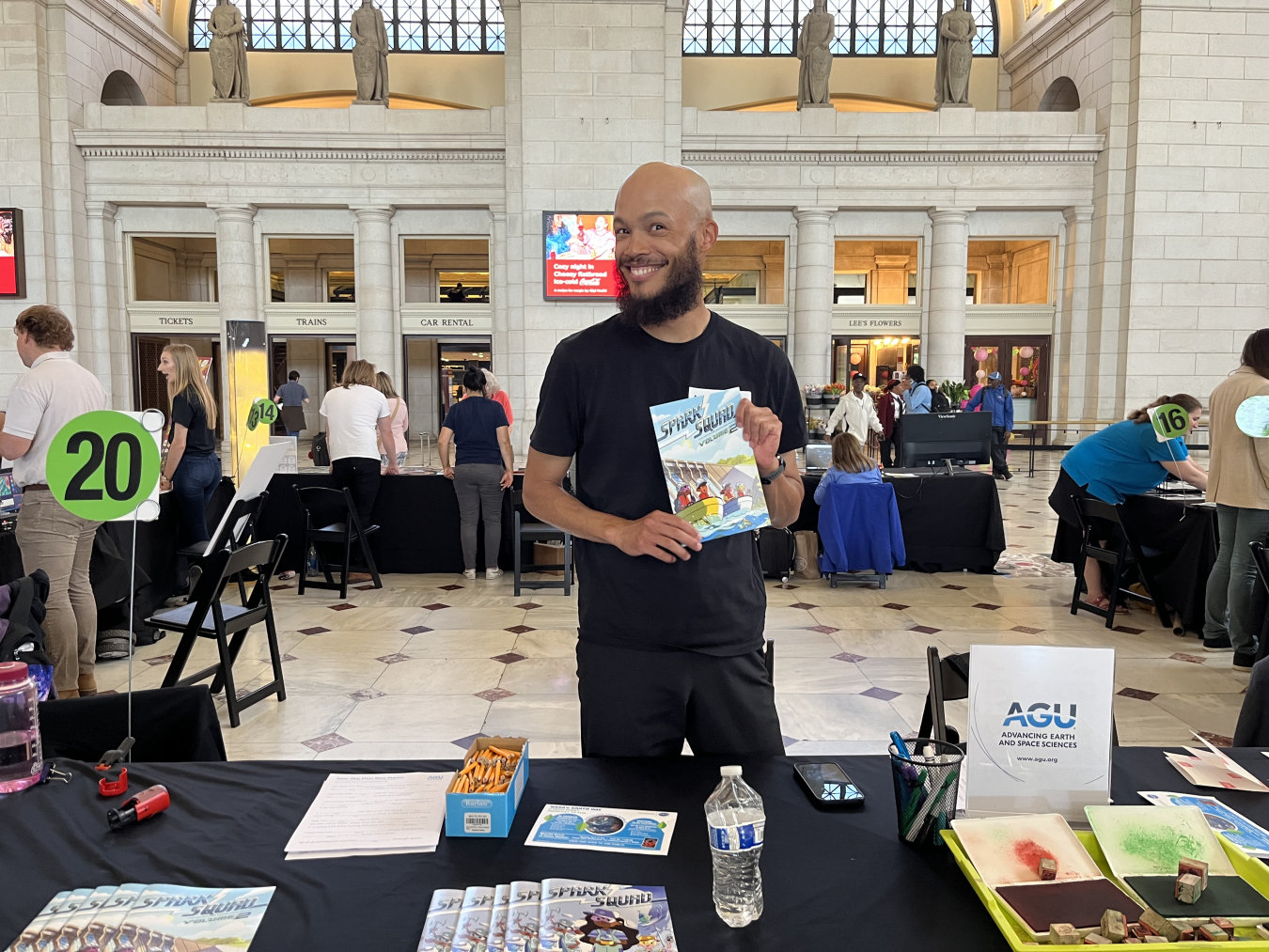 A person holding up copies of the Spark Squad while standing behind a table in a large room
