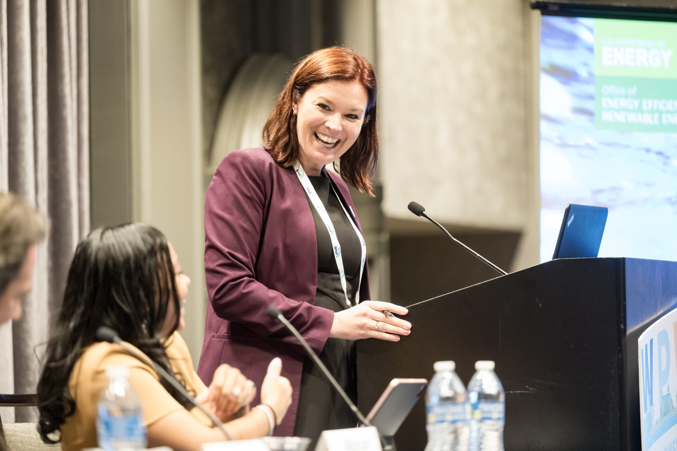 A woman standing at a podium smiles down at a panelist sitting at a table