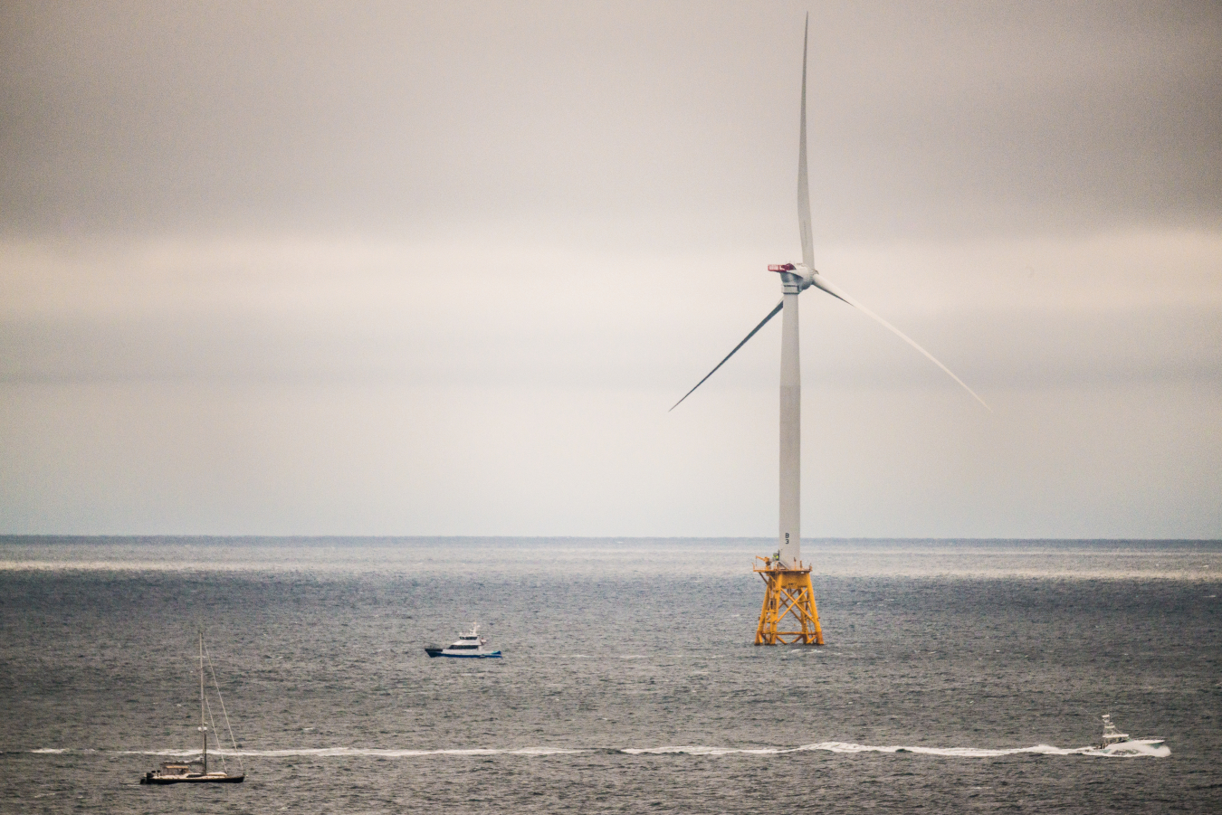 Boats sail around a large ocean-bound wind turbine