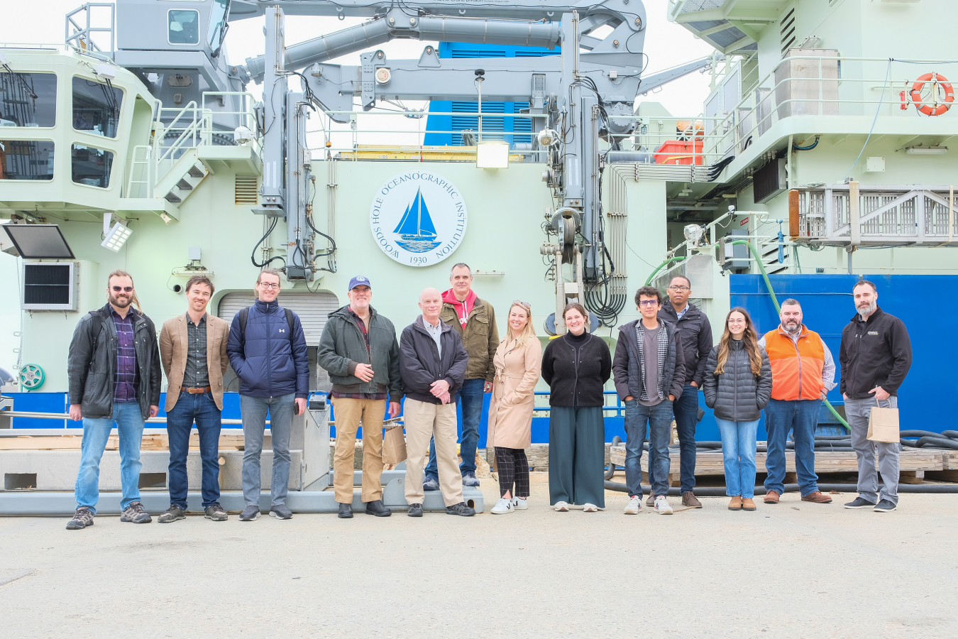 A group of people standing in front of an ocean research vessel as tall as a single-story building