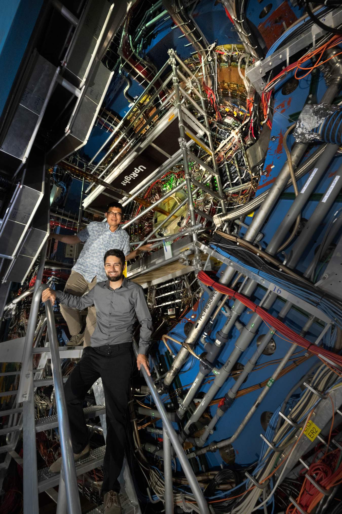 Researchers Daniel Brandenburg (a white man in a gray button-down shirt and black pants) and Zhangbu Xu (an East Asian man in a blue patterned button-down shirt and khaki pants) stand beside STAR, a house-sized machine with a giant blue ring in the front covered in wires and pipes.