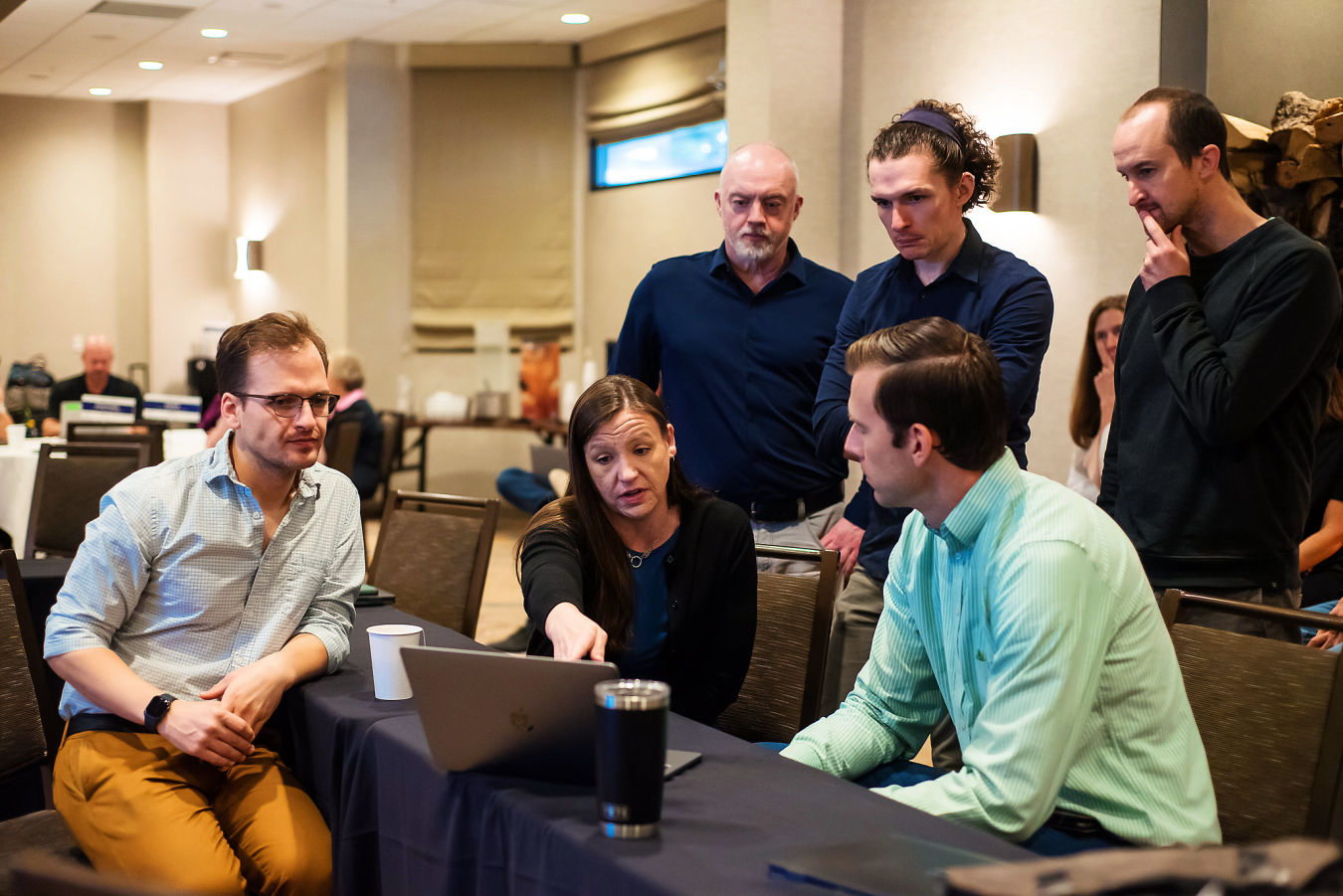 Three people sit at a table, looking and pointing at a laptop, while two people stand behind them.