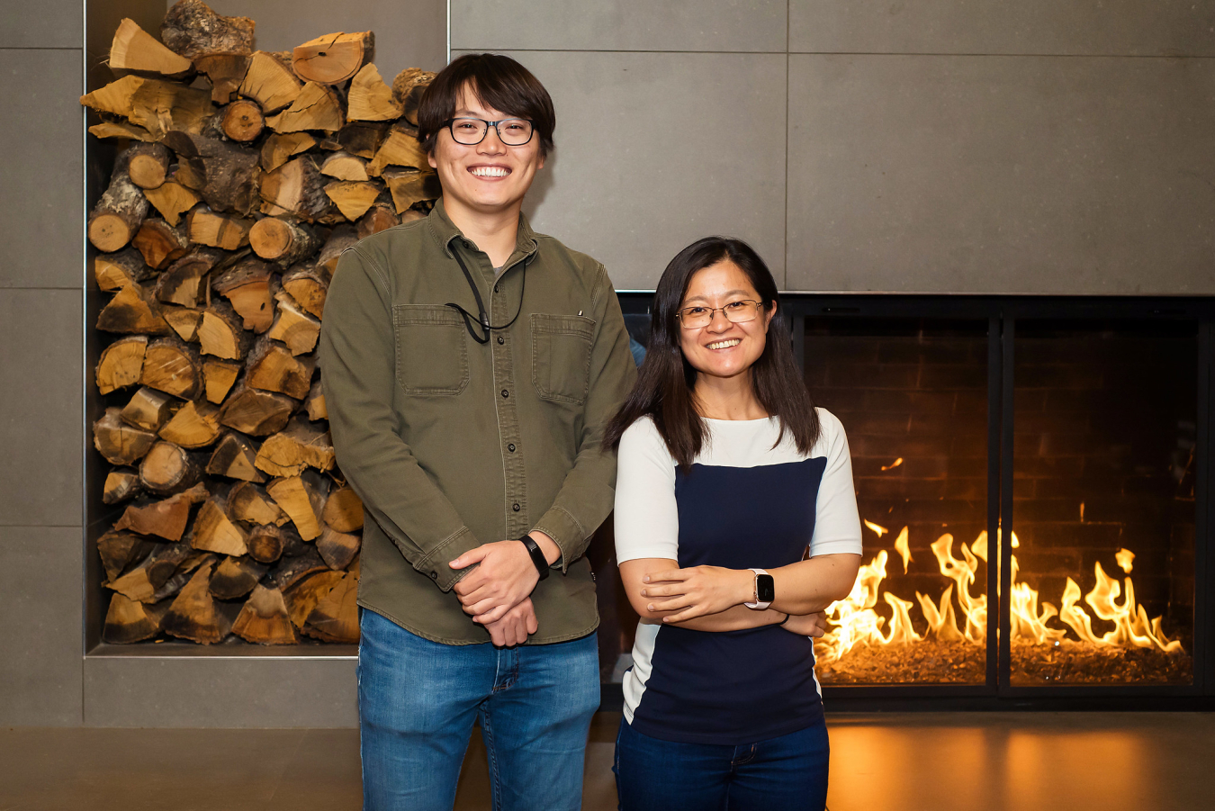 Two people pose and smile for the camera in front of an indoor fireplace.