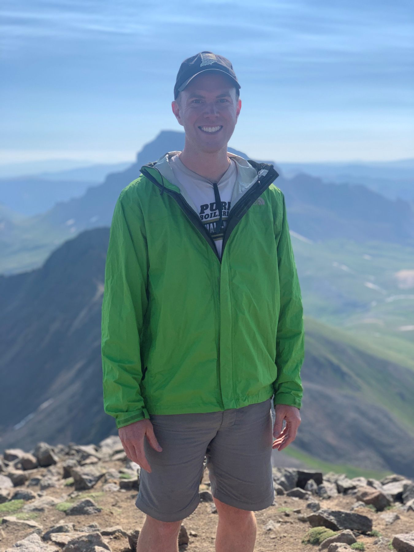 A man stands and smiles at the top of a mountain.