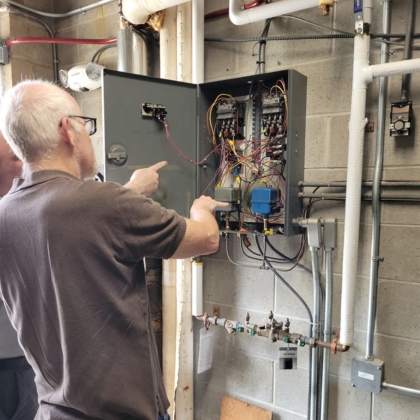 One man is captured pointing at an electrical box on the wall of a utility room. The hand of another man off-screen is also pointing at the box.