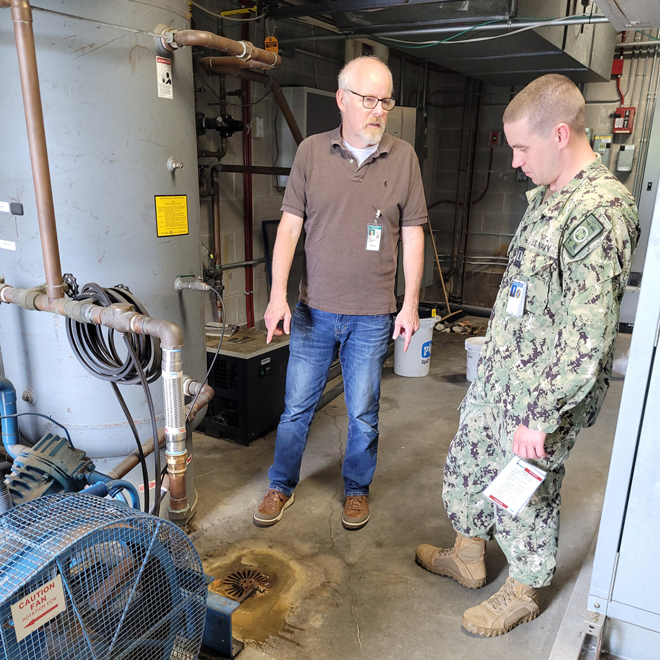 In a utility room, two men are grouped around mechanical equipment. One of them points to a pipe and is talking while the other looks at the area of interest.