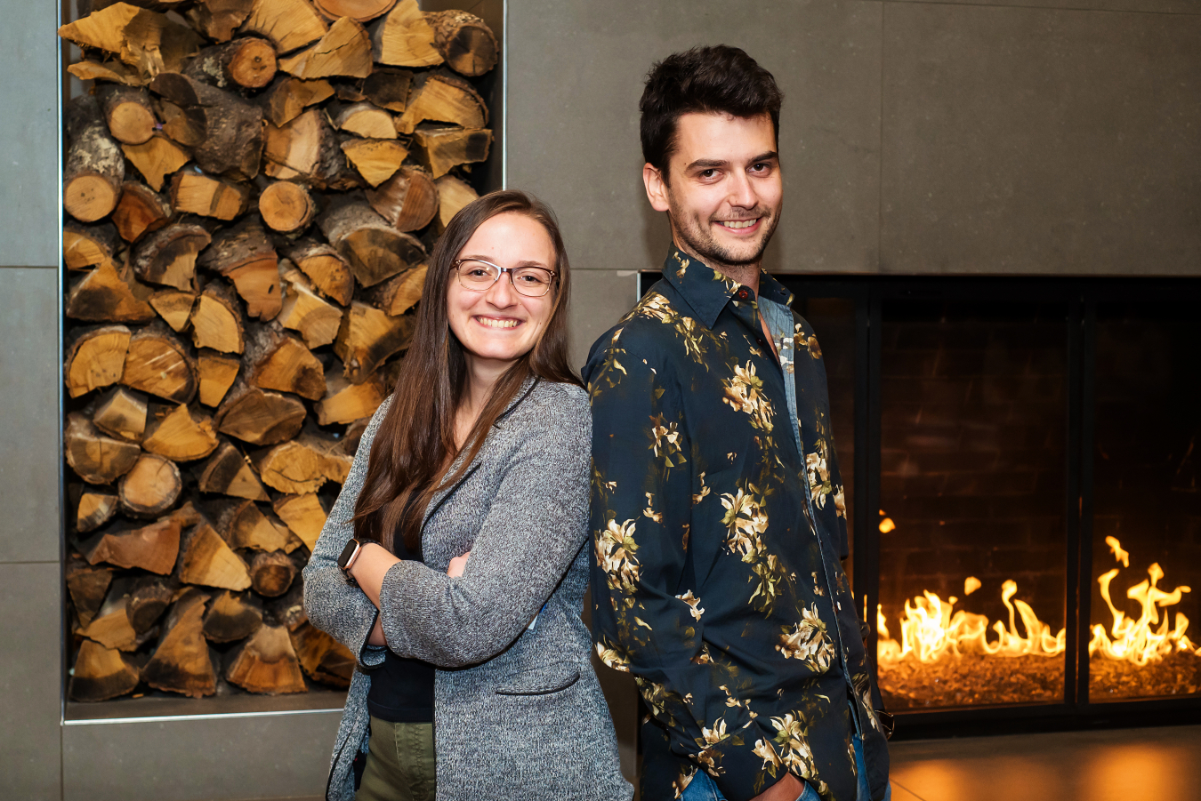 Two people pose back to back and smile for the camera in front of an indoor fireplace.
