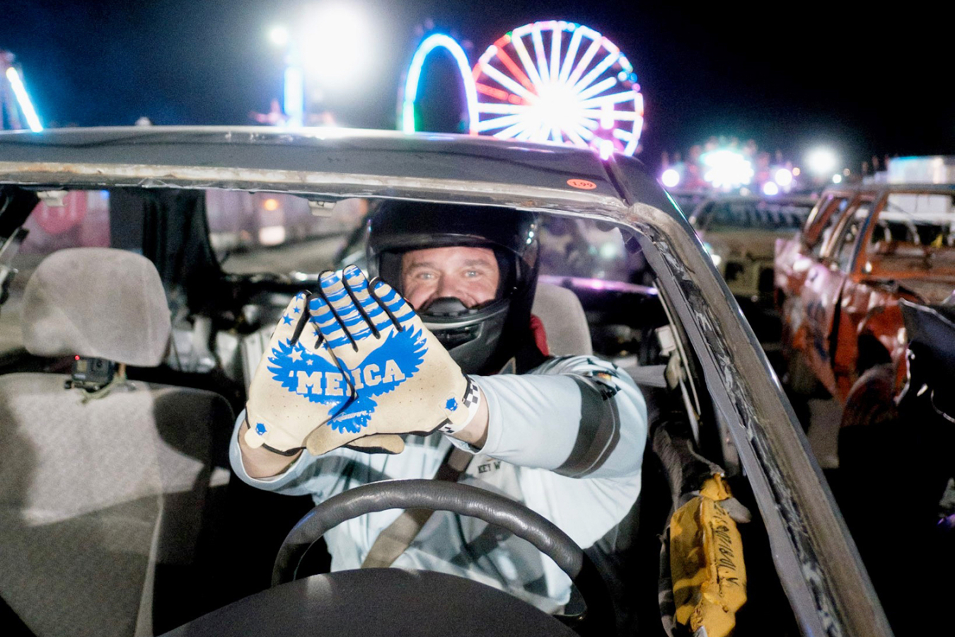 A driver at the wheel holds his gloves together to form the word 'Merica before the demolition derby race. Fairground rides are visible in the background.