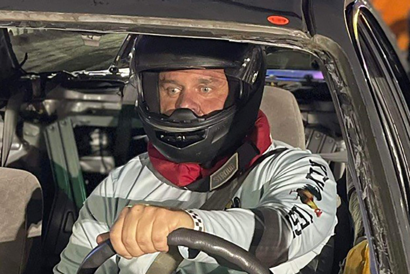 A man wearing a helmet sits at the wheel of a car staring.