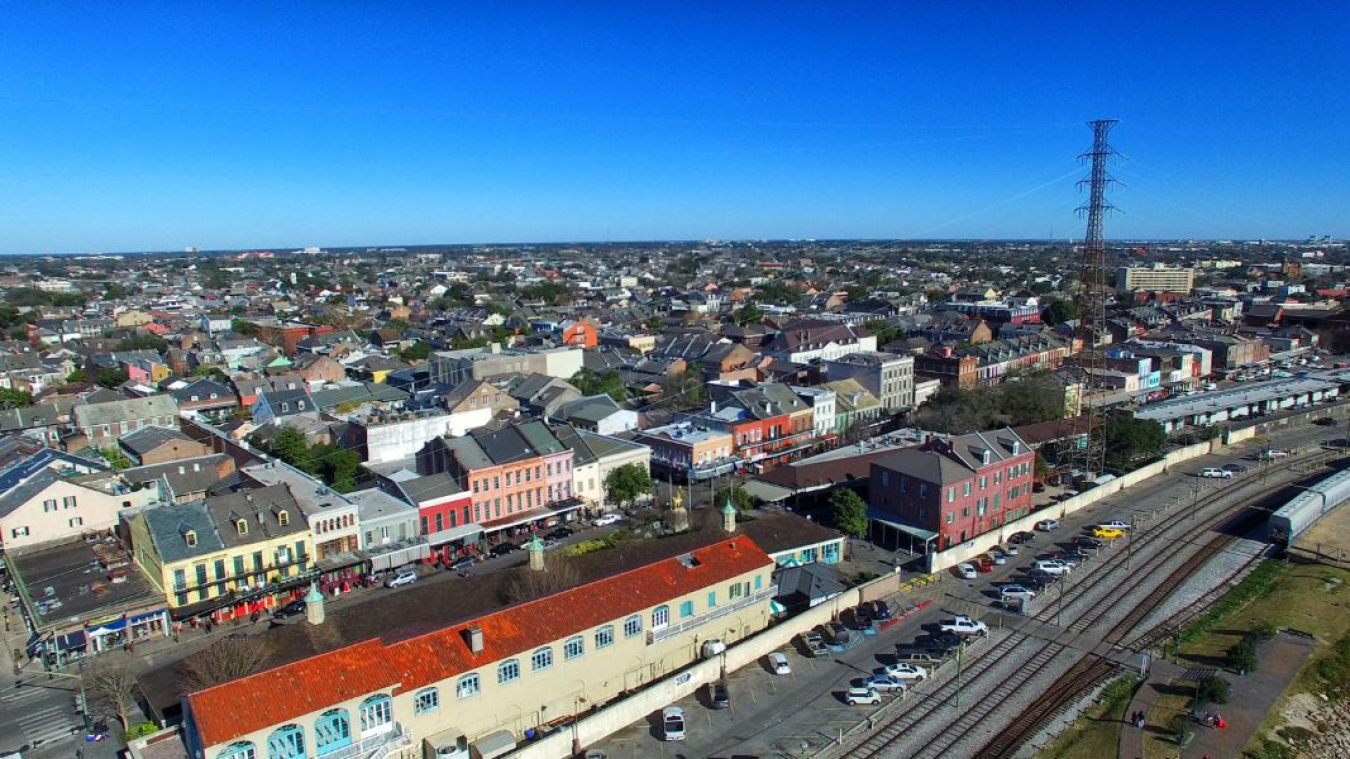 An aerial view of a compacted city landscape. There are train tracks, buildings, and houses surrounding a transmission tower and power lines in the foreground. In the background, is a cloudless, bright blue sky.