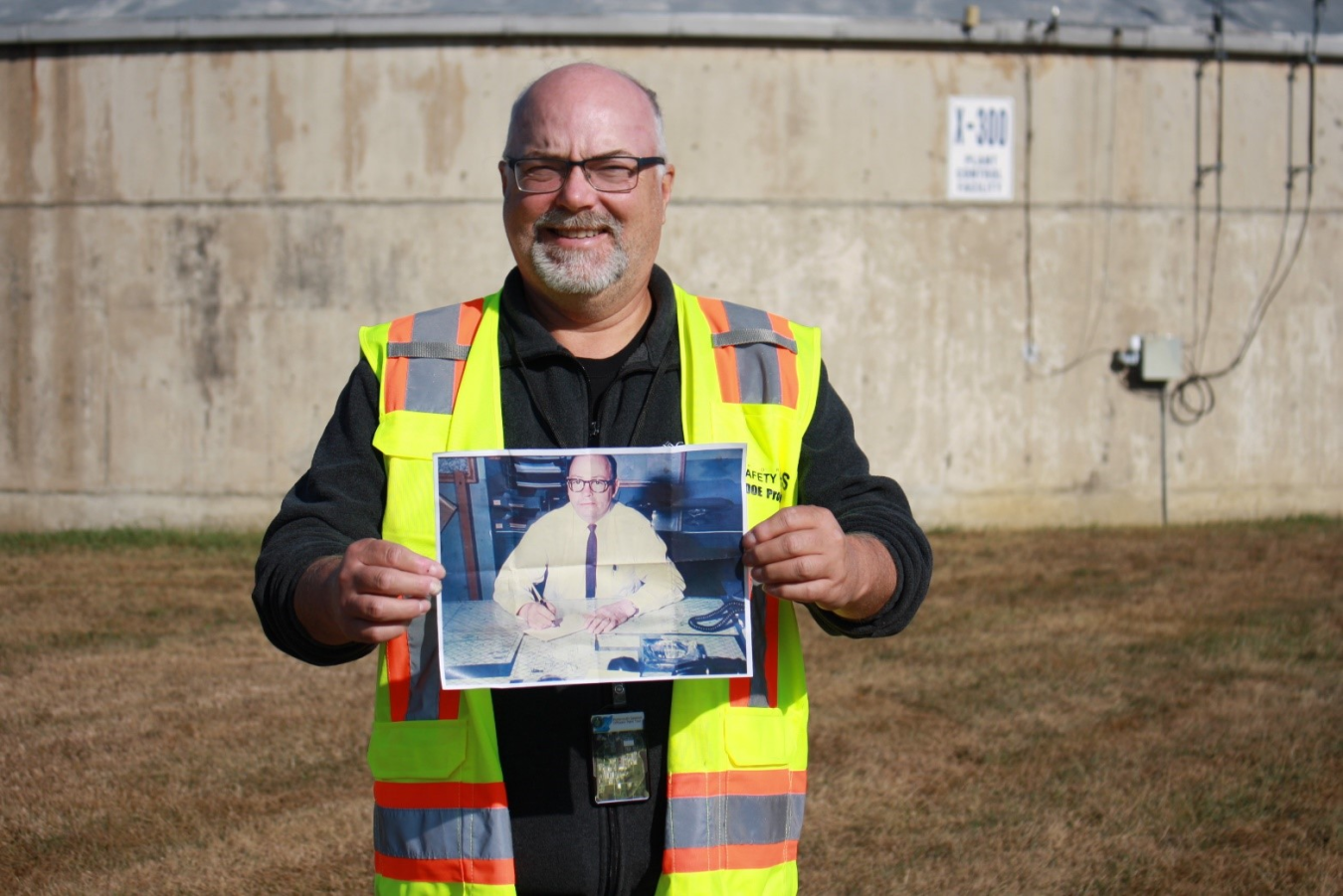 A man in a yellow safety vest holds up a photo of a man sitting at a table