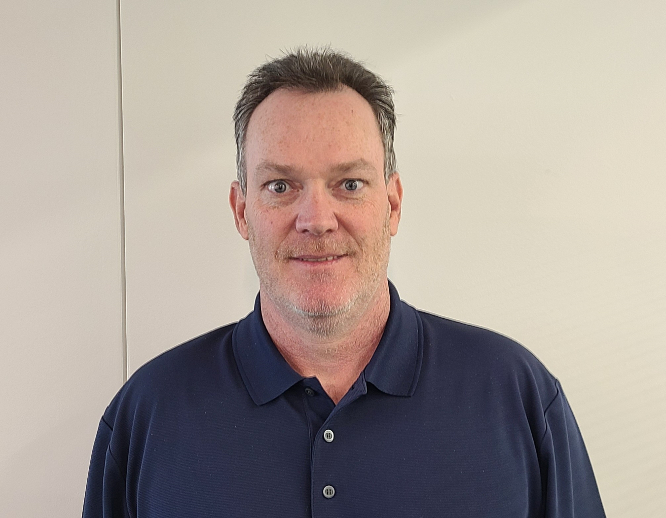 Man smiling with a blue collared shirt on in front of a beige wall