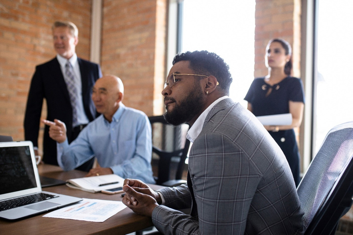 Four people of various ages sit and stand at a conference table looking out in the distance. 