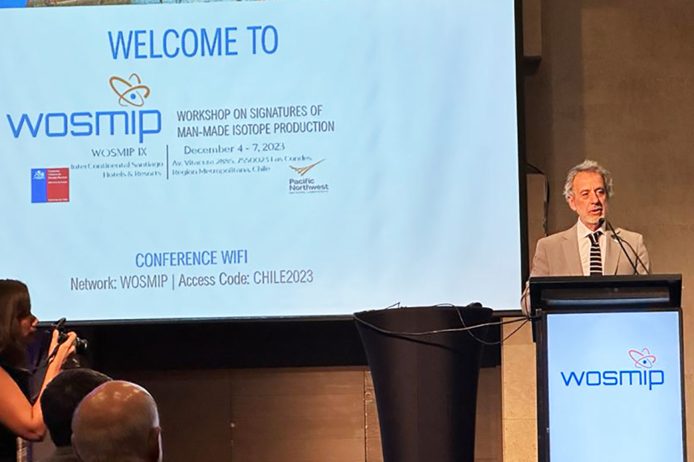 A man speaks at a podium. Words on a screen behind him and on the podium say WOSMIP.