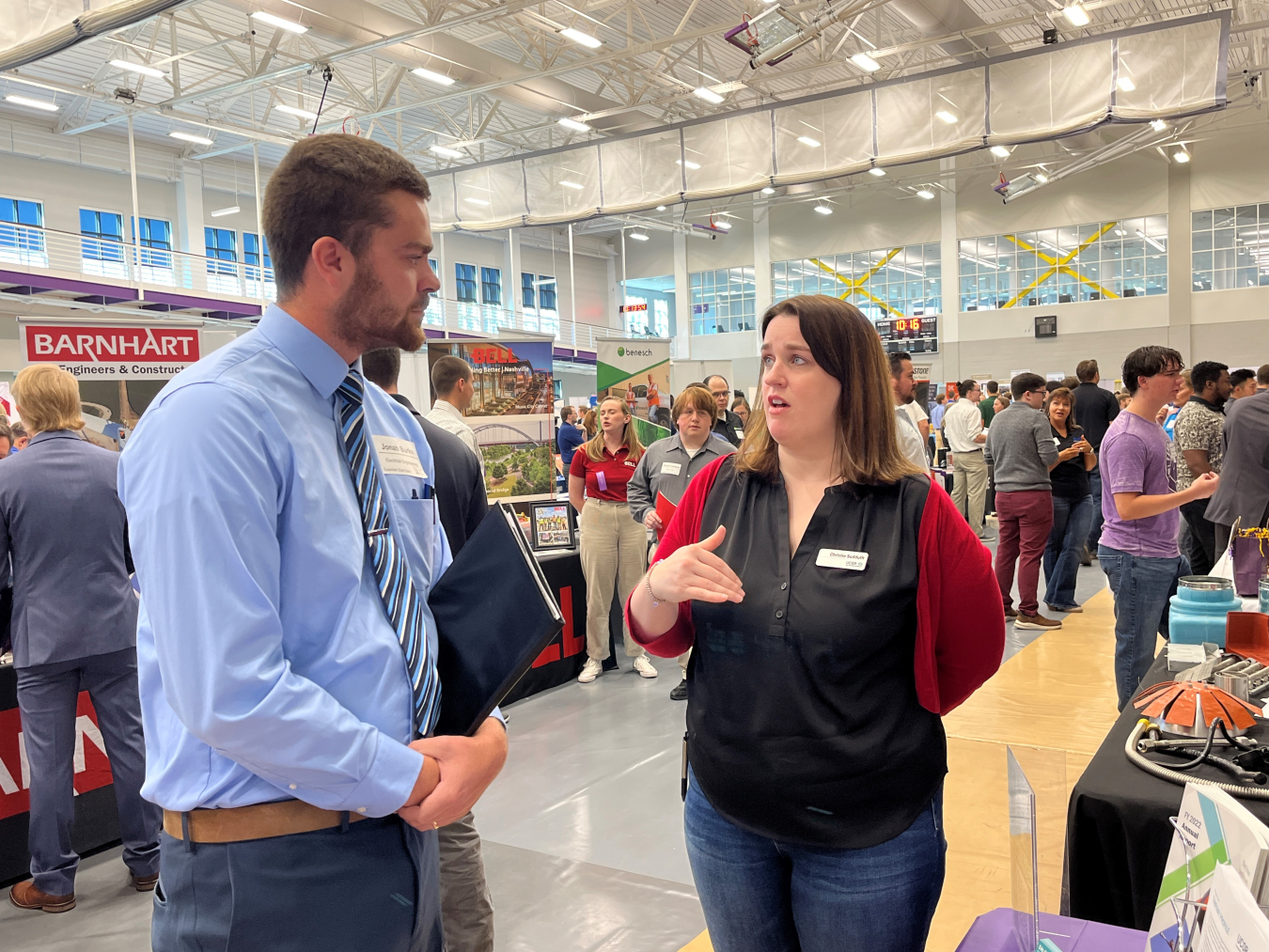 An employee stands at a booth and speaks to a student at a career fair