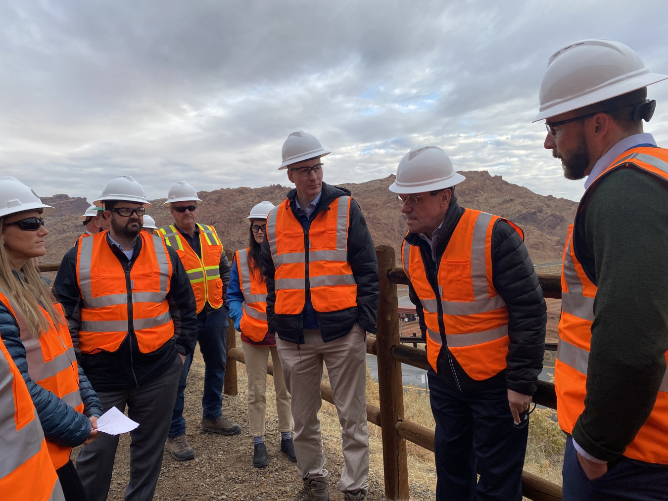 A group of people in orange safety vests and white hard hats stand and talk
