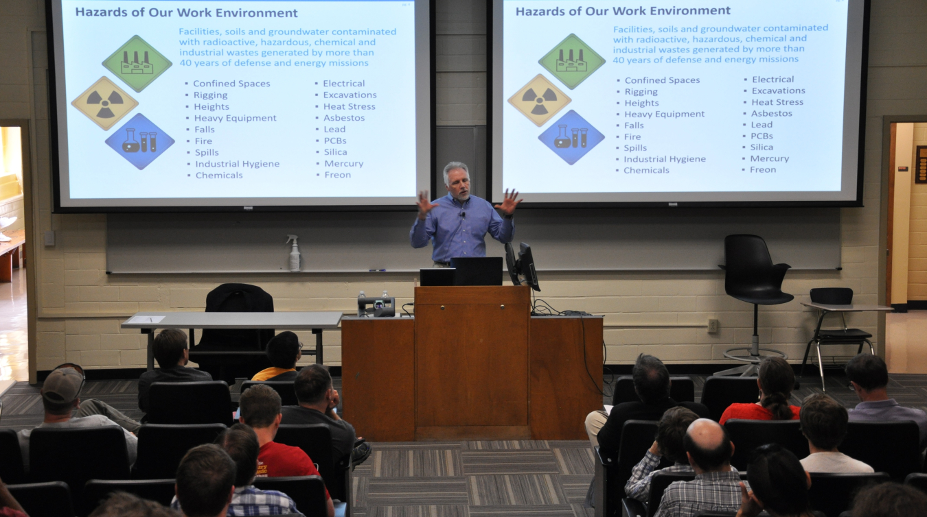 A man stands behind a desk and lectures to a college class