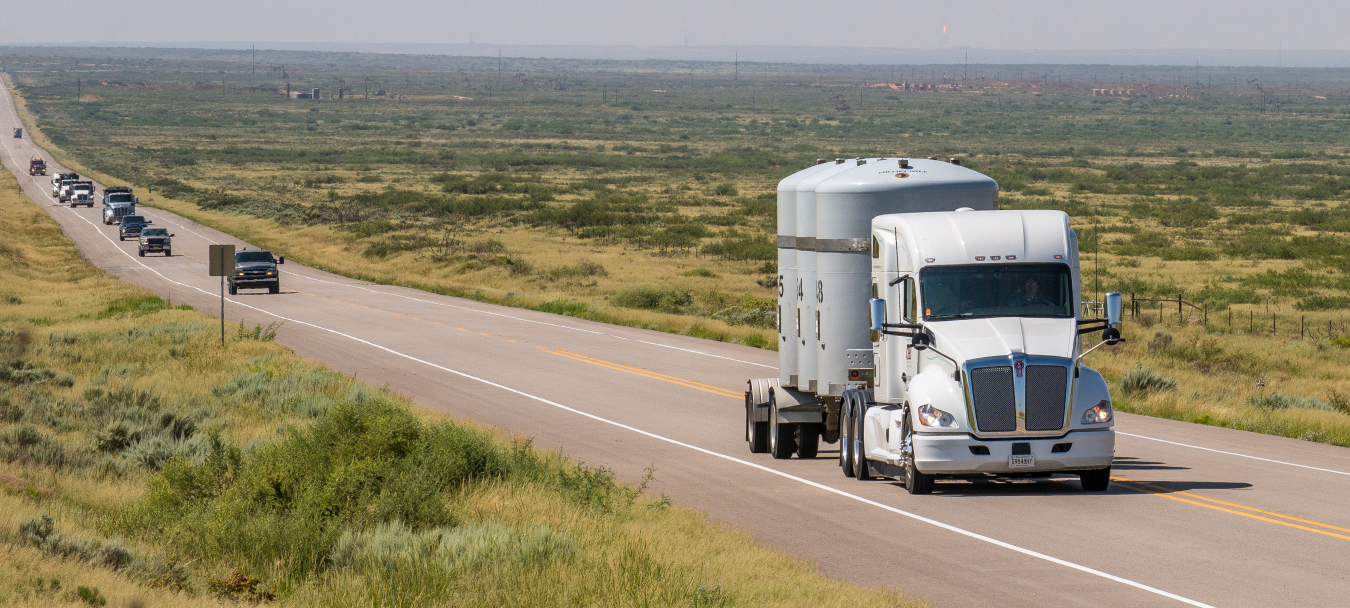 A large white tractor trailer truck drives on a road with cars behind it 