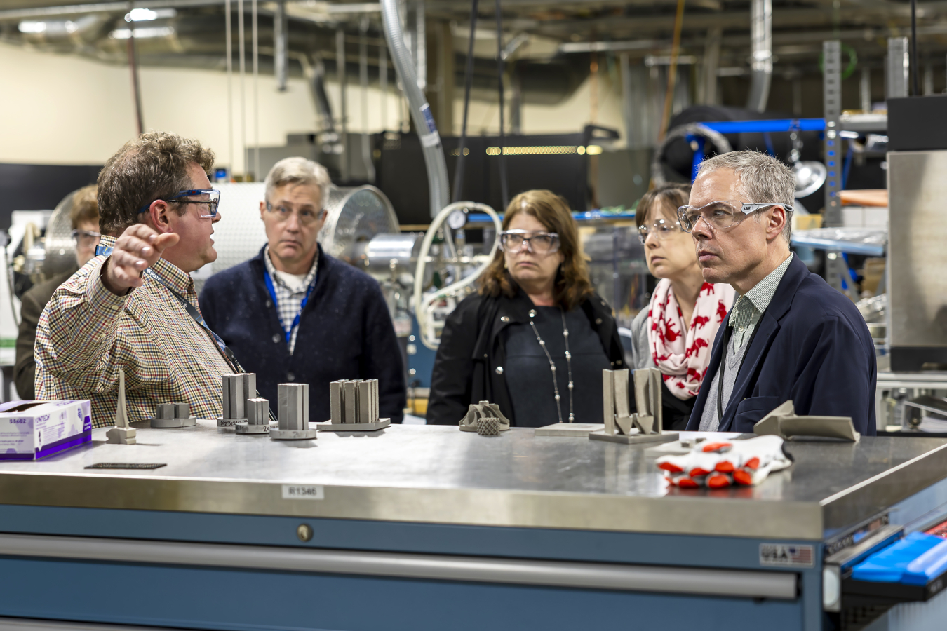 Man stands in front of metal workbench with hand out speaking to group of visitors at a facility.