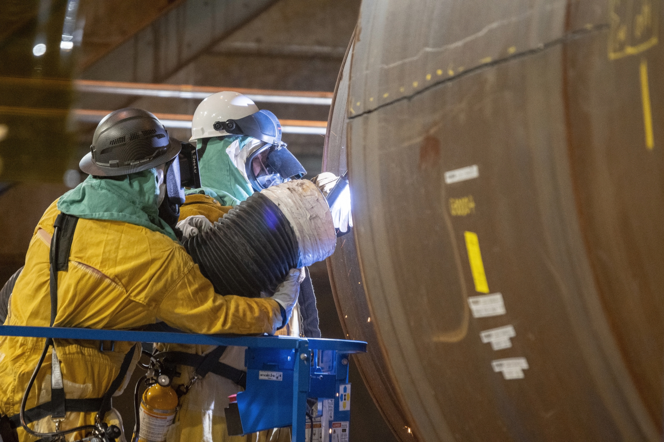Workers using tools on a large pipe while dressed in hazmat suits