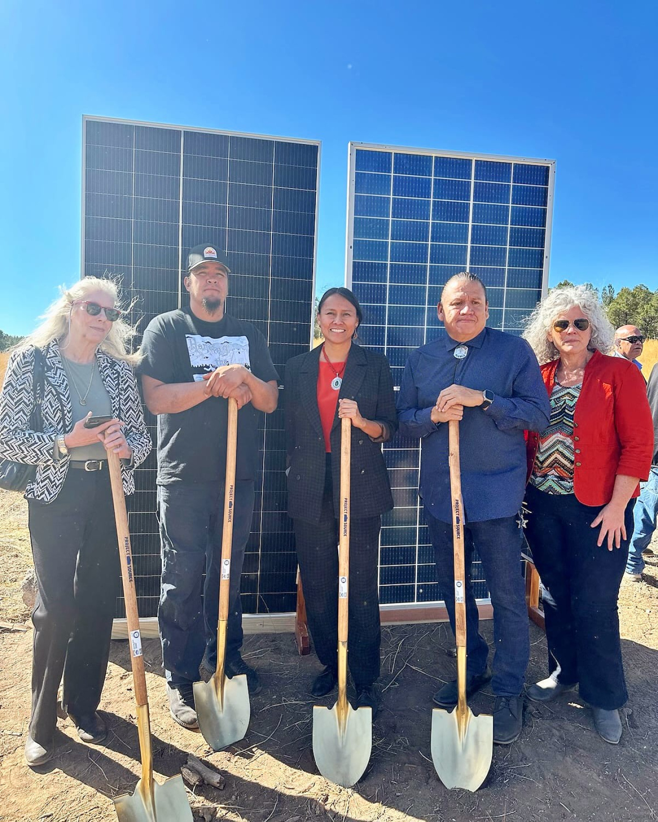 Two men and three woman, all with shovels, pose for a photo with a solar panel in the background.