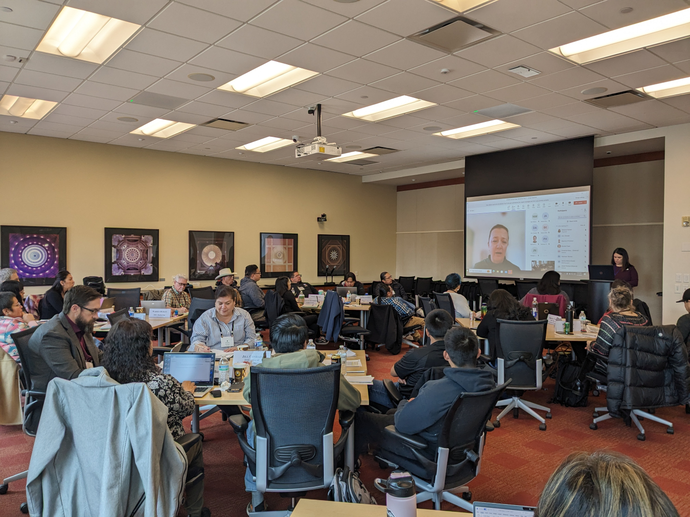 A group of people in a large classroom watch a presentation on a screen 