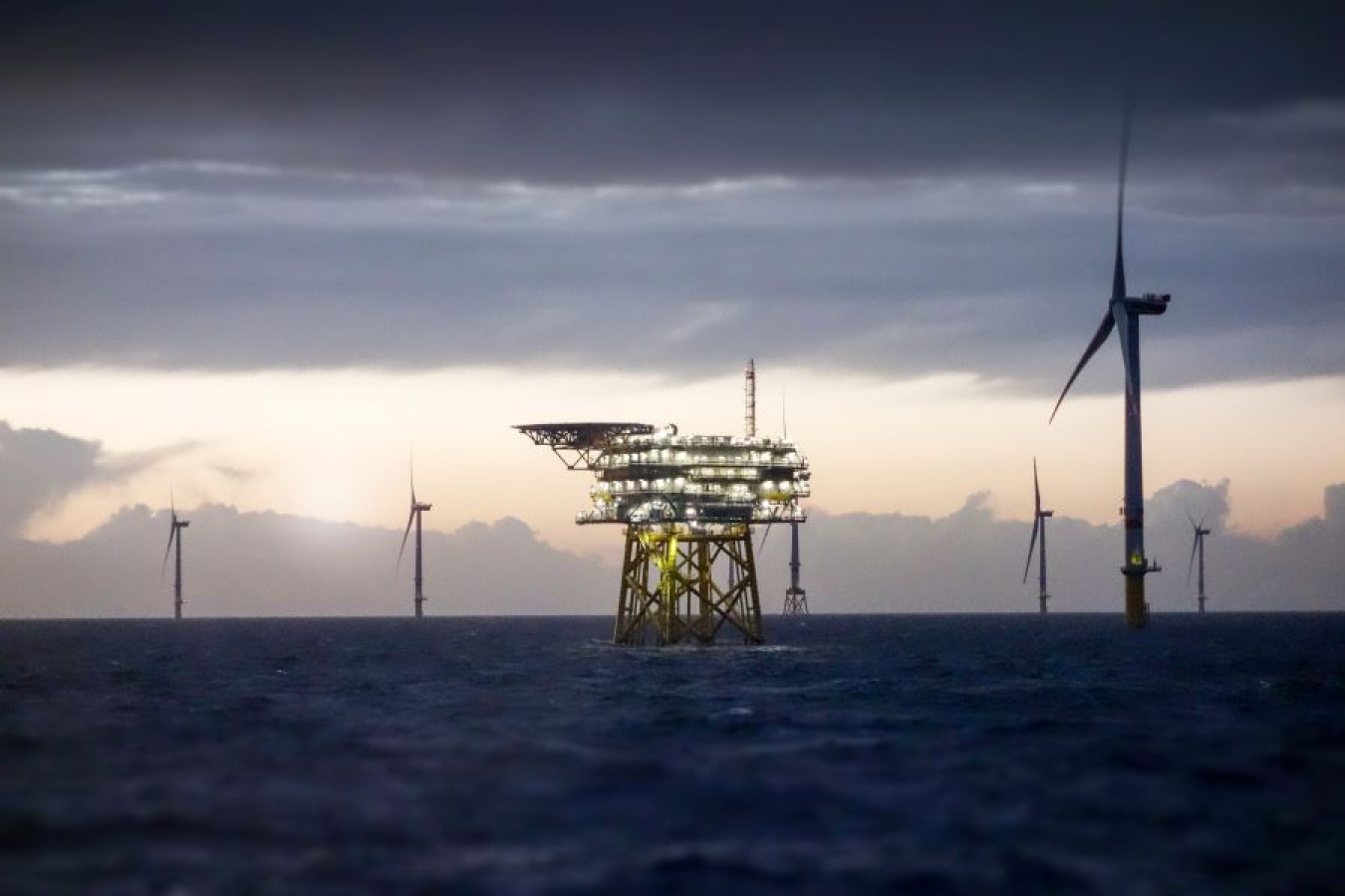 An offshore platform substation and wind turbine farm sits in the middle of a dark sea with a cloudy sky in the background.