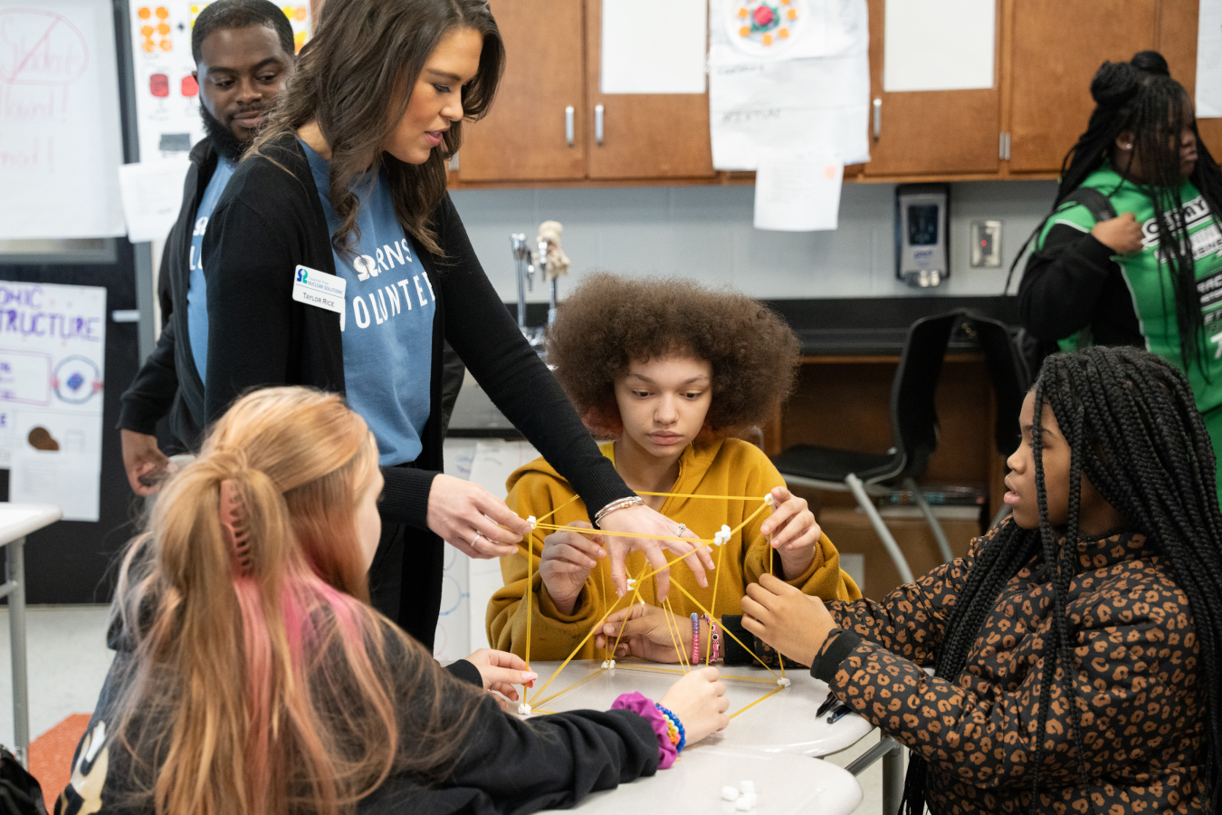 Students sit at a table while an engineer stands and helps them build a structure made from spaghetti and marshmallows.