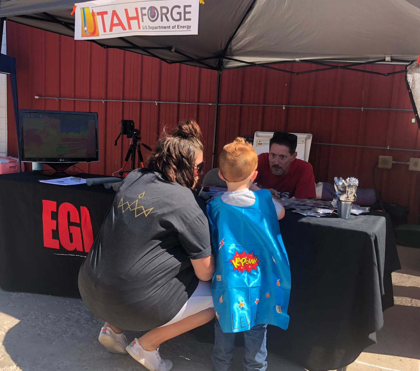 A small child in a cape and his guardian stand in front of the Utah FORGE booth, where Christopher Katis answers questions about geothermal energy.