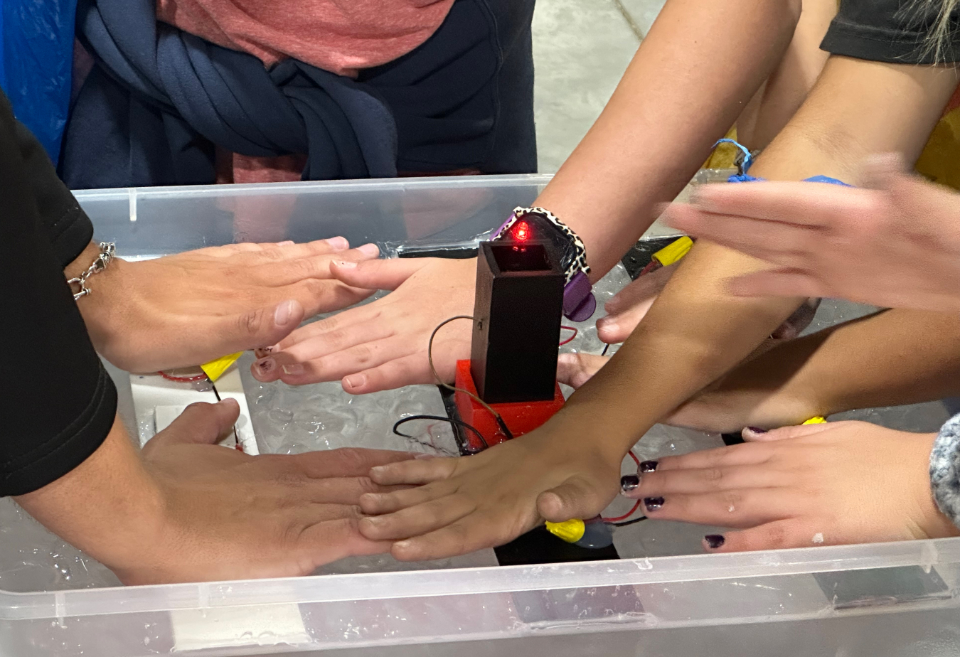 Several peoples hands' press down flat on a Peltier Module, which has three planks with electrodes and wires connecting them lying on a bed of ice water.