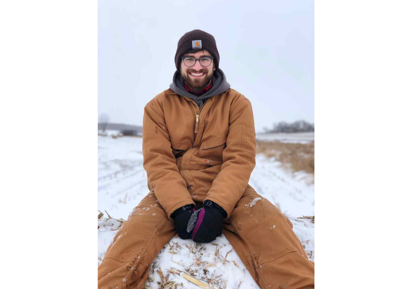 Augustus Pendleton wearing coveralls sitting atop a snow-covered bale of corn stover