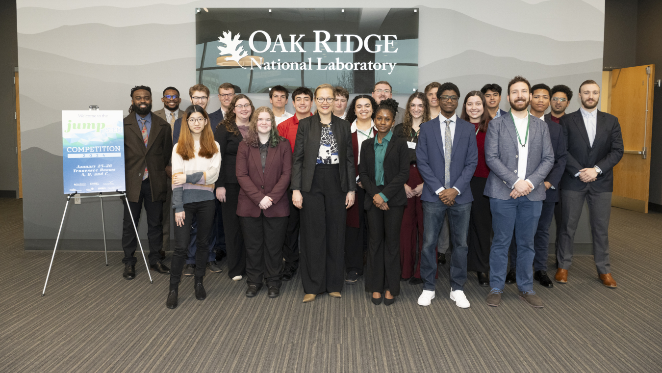 A group of people standing and facing the camera, with the sign "Oak Ridge National Laboratory" behind them, and a smaller presentation sign on a stand next to them.