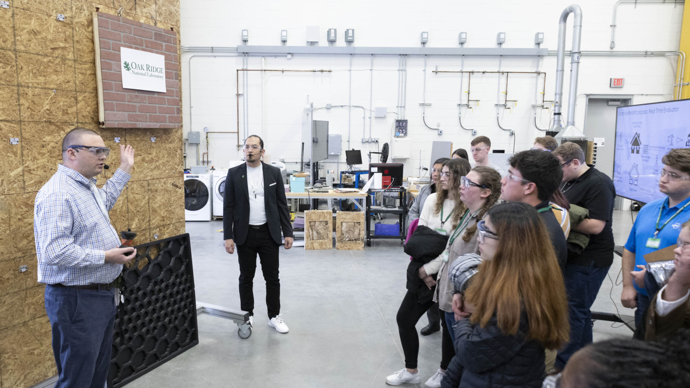 A man standing near a wall in a laboratory workroom, talking to a group of people standing nearby.