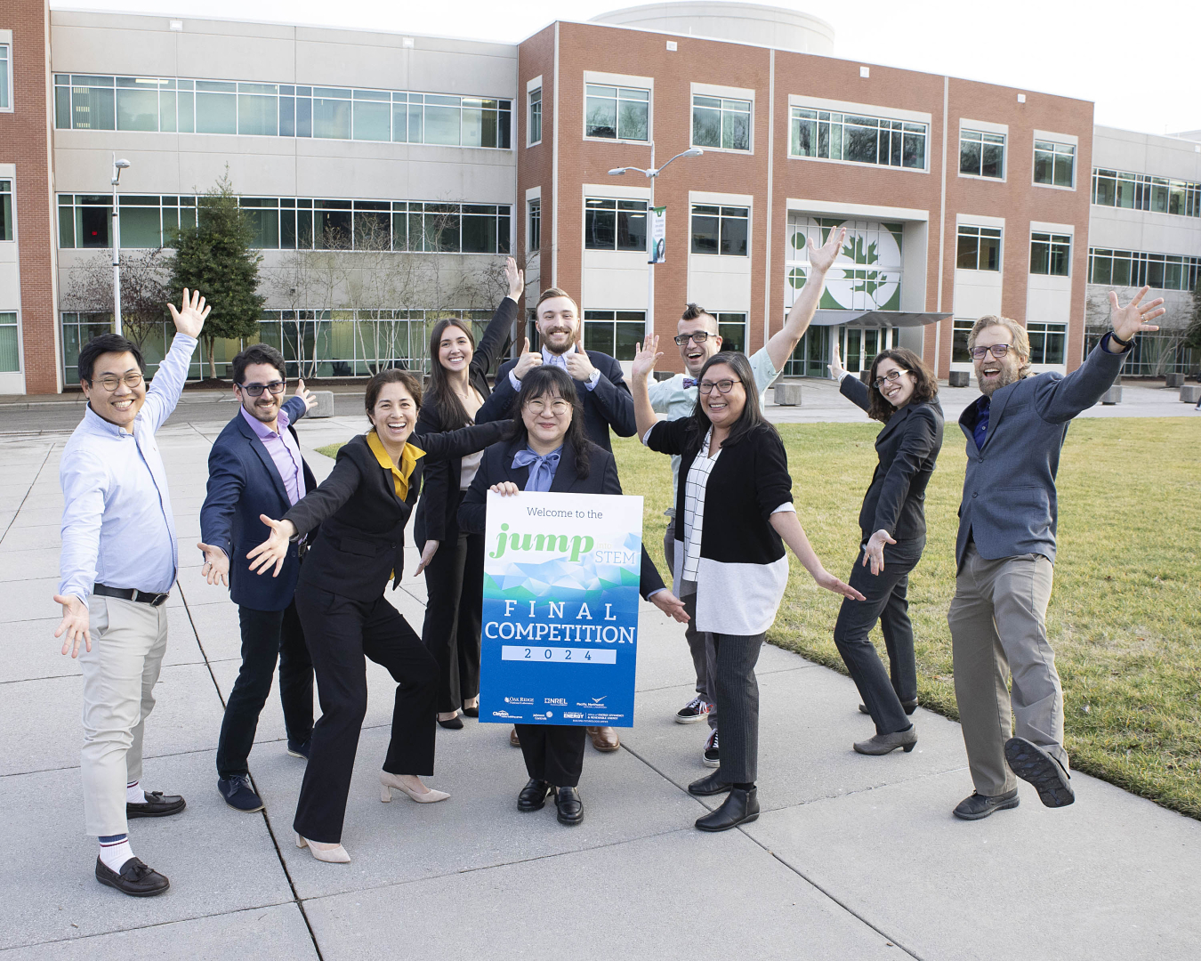A group of people standing outside the Oak Ridge National Laboratory building, some of them with their arms raised exuberantly and some holding a poster sign..