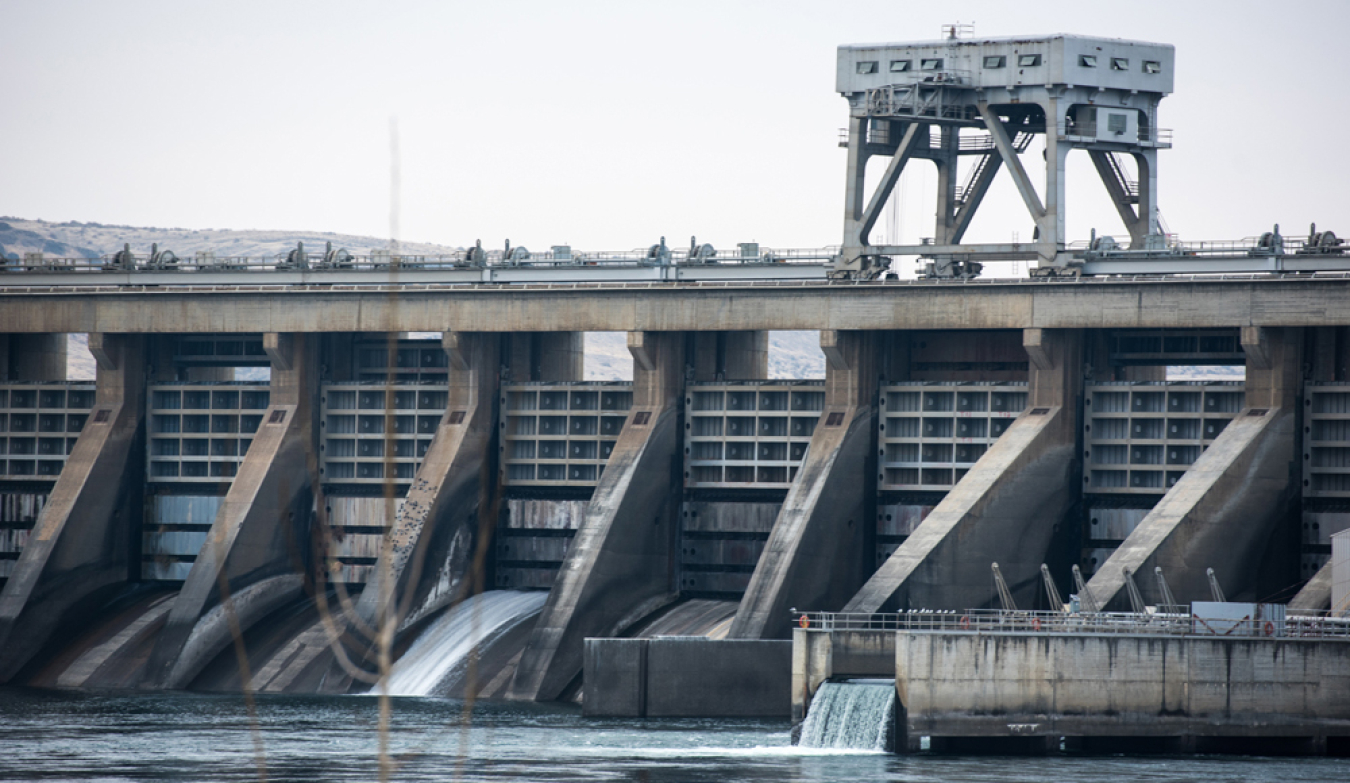 A view of part of the McNary Dam in Umatilla, Oregon, on a gray, overcast day. Water can be seen pouring out of two spillways and into the reservoir of water in the foreground of the photo. 