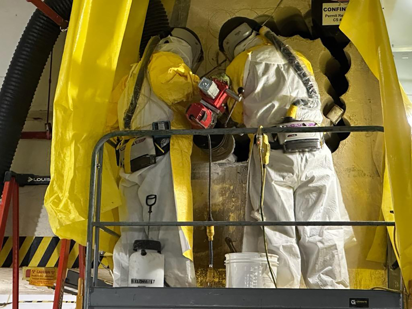 Workers wearing white hazmat suits use large drills and tools on an air duct