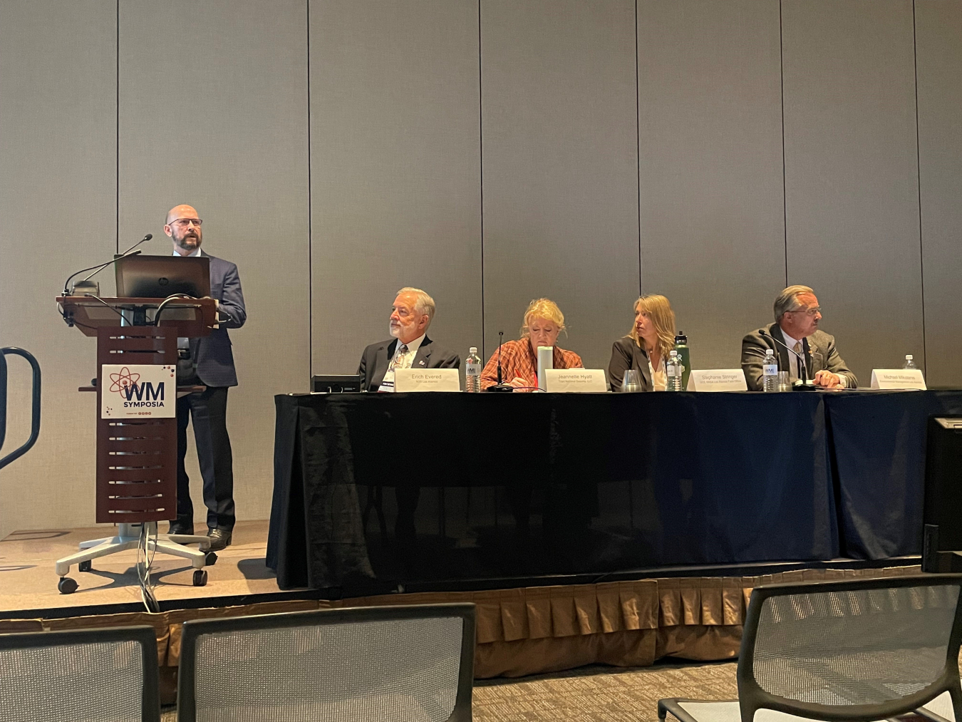 A man standing at a podium speaking beside a table of panelists listening to him